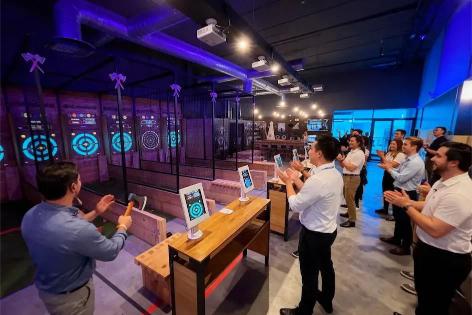 Un grupo de personas en traje de negocios de pie y aplaudiendo mientras otros lanzan hachas a objetivos en un lugar de lanzamiento de hachas de interior, perfecto para Team Building Las Rozas.